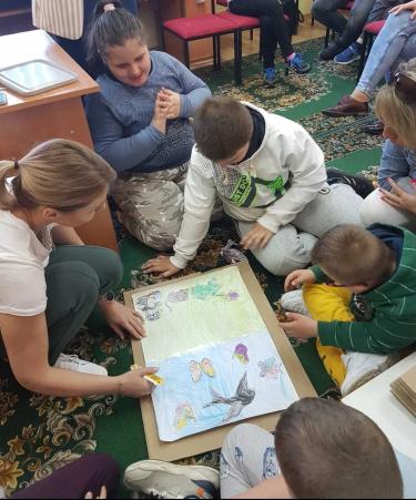 Uczniowie w raz z nauczycielką siedzą na podłodze i przyglądają się leżącym na niej kolorowej kartce i  doklejają do nich wycięte ilustracje.
