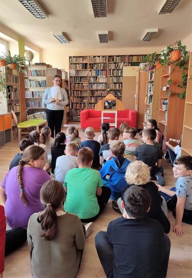 Zajęcia biblioterapeutyczne w SP Nr 1 w Przeworsku - dzieci siedzą w grupie na podłodze tyłem do fotografa, w tle stoi nauczycielka prowadząca zajęcia, widać rozłożony teatrzyk kamishibai i regały biblioteczne z książkami.