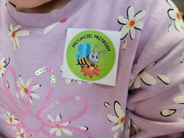 Widać część bluzy fioletowej w kwiatki a na niej przyklejona karteczka z zielonym napisem "Przyjaciel przyrody", pod napisem namalowana pszczółka uśmiechnięta.