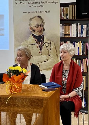 Panie Stanisława Żak i Lidia Kiełczyńska siedzą za stolikiem i słuchają przemówień przybyłych gości.